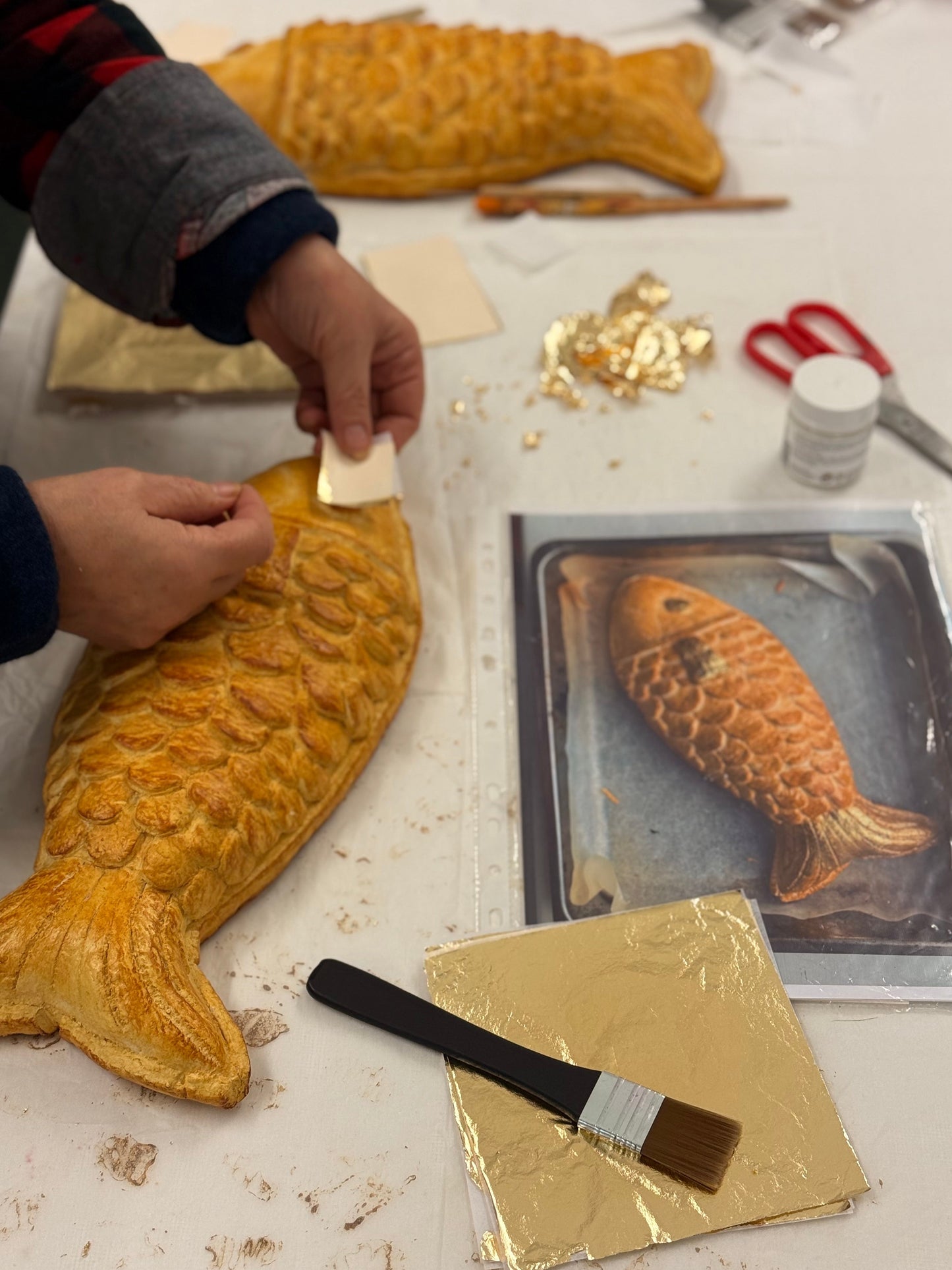 Person decorating a fish-shaped pastry with gold leaf on a table with tools and gold foil.